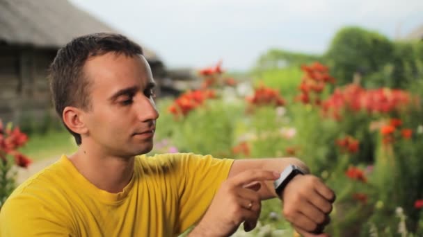 Hombre joven tocando reloj inteligente cerca de flores en el jardín. Comprueba los mensajes con el telón de fondo de flores blancas, rojas y púrpuras — Vídeos de Stock