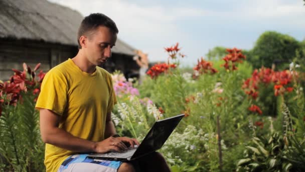 Jovem trabalhando em um laptop perto de camas de flores. Flores brancas, vermelhas e roxas no fundo — Vídeo de Stock