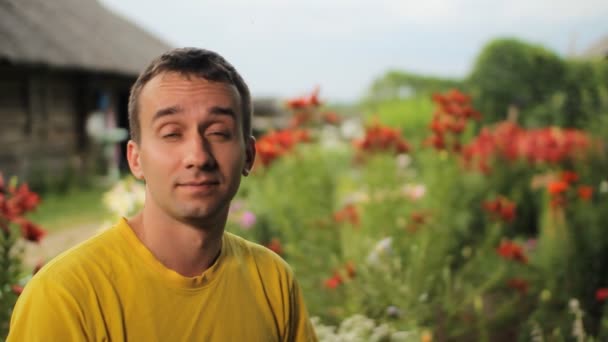 Молодий чоловік посміхається біля квітів у саду. На тлі білих, червоних, фіолетових квітів — стокове відео