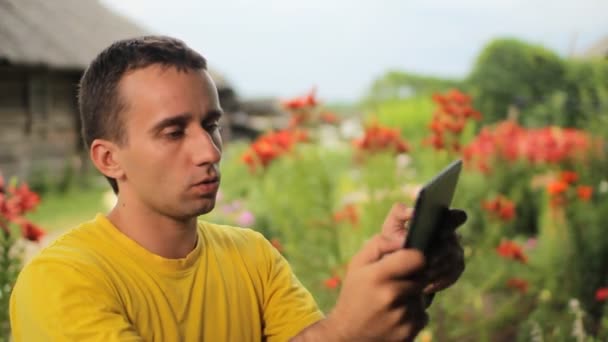 Hombre joven utilizar la tableta para el juego cerca de flores en el jardín. Flores blancas, rojas y moradas en el fondo — Vídeos de Stock