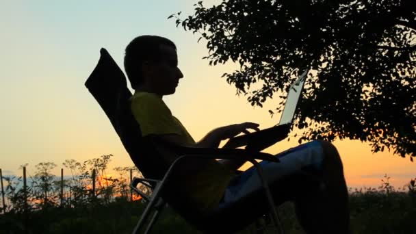 Slhouette de hombre de negocios feliz con el ordenador portátil trabajando en el campo al atardecer. Hombre balanceándose en silla — Vídeos de Stock