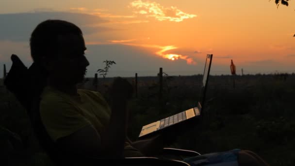 Slhouette de hombre de negocios feliz con el ordenador portátil trabajando en el campo al atardecer. Hombre balanceándose en silla — Vídeos de Stock