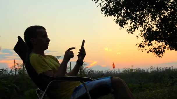 Un hombre se sienta en una silla con el teléfono. Sobre el fondo de un cielo anaranjado después del atardecer — Vídeos de Stock