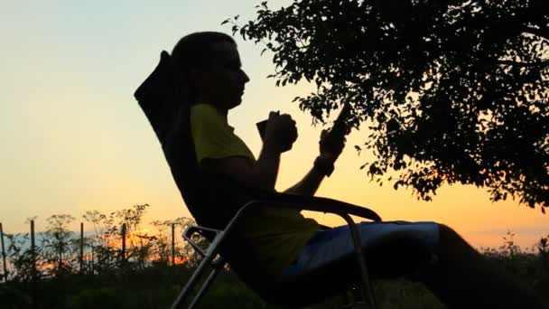 Um homem senta-se numa cadeira com o telefone e uma chávena de chá. Contra o pano de fundo de um céu laranja após o pôr do sol — Vídeo de Stock