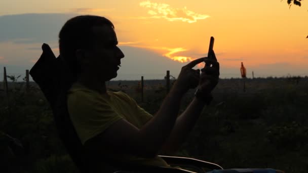 En man sitter i en stol med telefonen. Mot bakgrund av en orange himlen efter solnedgången — Stockvideo