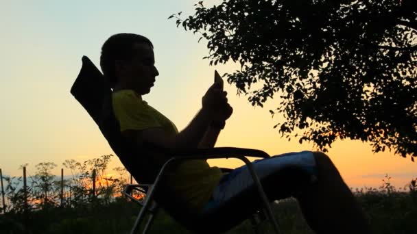 Силуэт мужчины в кресле играет на планшете. На фоне оранжевого неба после заката — стоковое видео