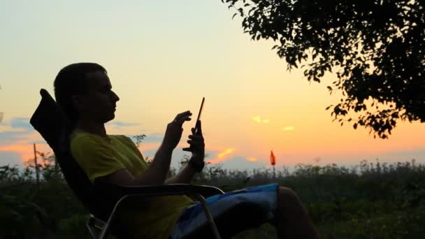 Silueta de hombre en silla jugando en tableta. Sobre el fondo de un cielo anaranjado después del atardecer — Vídeos de Stock