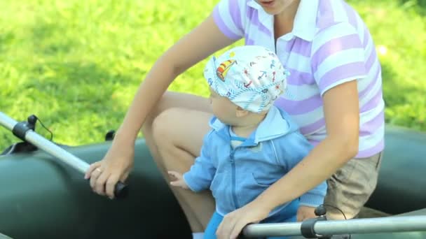 Vackra pojke leker med en uppblåsbar båt och en paddel med mamma. Båten är på gräset i trädgården. Växande framtida fiskare — Stockvideo