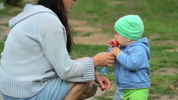 息子は、ボトル入り飲料水と彼女の腕の中で彼を保持している母親を保持しています。庭で屋外撮影. — ストック動画