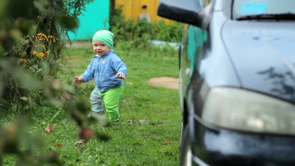 Hermoso niño caminando en el jardín cerca de la casa. Cerca hay un coche oscuro y las manzanas crecen. Niño con sombrero y chaqueta — Vídeo de stock