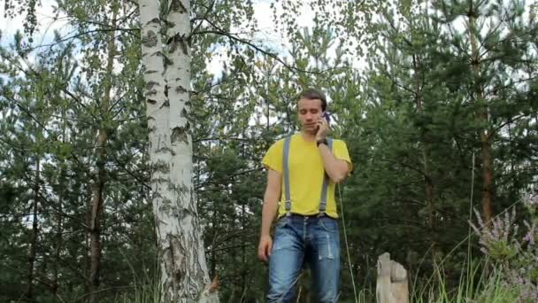 Man wandelaar wandelen door het bos en praten op de telefoon. Geel T-shirt en jeans met bretels. Technologie in het wild — Stockvideo
