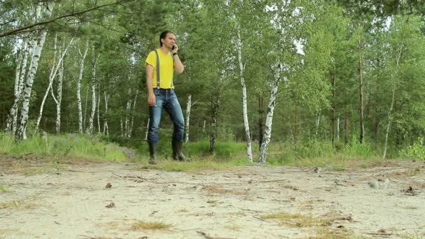 Un escursionista che cammina nella foresta e parla al telefono. T-shirt gialla e jeans con bretelle. Tecnologia in natura — Video Stock