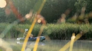 Balıkçı şişme bir teknede nehirüzerinde yüzer. Güneş ışınları çerçevede oynar. Vahşi doğa aktiviteleri
