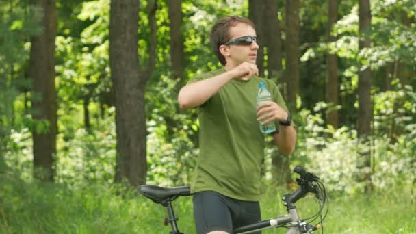 Unga manliga cyklist dricker vatten ur en flaska i parken. Grön T-shirt och solglasögon — Stockvideo