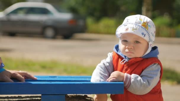 A boy stands near the bench on the playground with the honest expression. The boy is less than a year, a red vest and a white hat — Stock Video