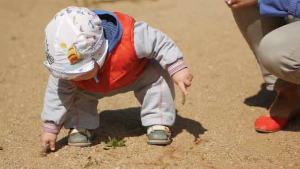 Um miúdo de um ano a brincar com areia no parque infantil. Perto da mãe ajuda-o com o jogo. Menino de colete vermelho e uma cobertura para a cabeça branca — Vídeo de Stock