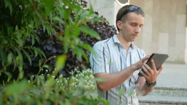Dijital tablet ile modern genç adam açık portresi. Güneş gözlüğü ve ceket bir adam. Yeşil bitkiler bir sürü