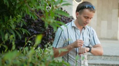 Modern genç adam akıllı saati olan açık portresi. Güneş gözlüğü ve ceket bir adam. Yeşil bitkiler bir sürü