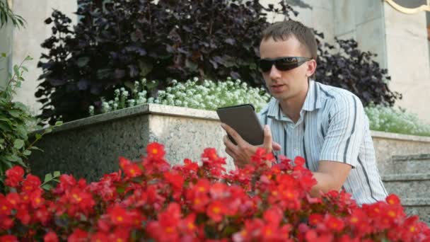 Retrato al aire libre del joven moderno con tableta digital. Hombre con gafas de sol y chaqueta. Cerca de muchas flores rojas — Vídeos de Stock