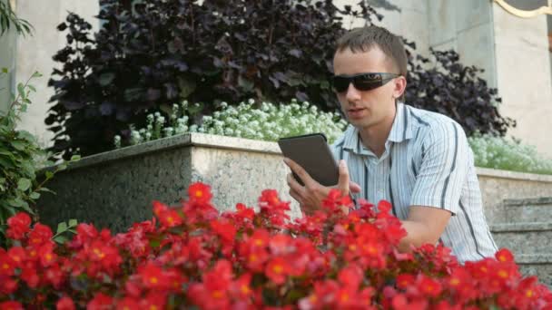 Retrato al aire libre del joven moderno con tableta digital. Hombre con gafas de sol y chaqueta. Cerca de muchas flores rojas — Vídeo de stock