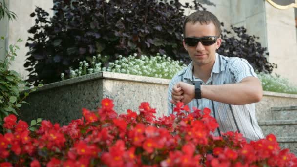 Retrato ao ar livre do jovem moderno com relógio inteligente. Homem de óculos de sol e casaco. Perto de um monte de flores vermelhas — Vídeo de Stock