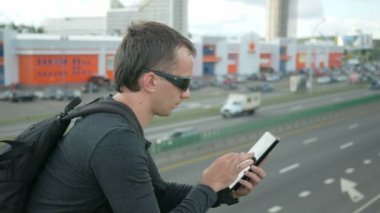 Modern genç adam sokakta dijital tablet ile açık portresi. Sırt çantası köprüde oturan gözlüklü adam. Alt araba sürmek