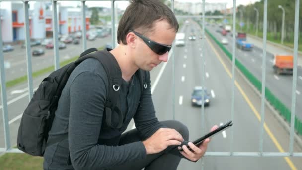 Portrait extérieur de jeune homme moderne avec tablette numérique dans la rue. L'homme en lunettes avec un sac à dos assis sur le pont. Voitures de fond — Video