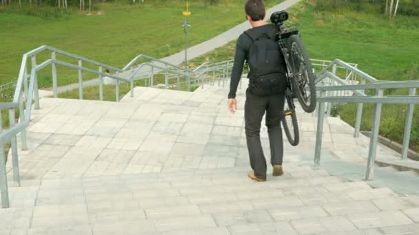 Un joven lleva una bicicleta a través de las largas escaleras. En el fondo del campo verde. Nubes cambiando escaleras de luz — Vídeo de stock