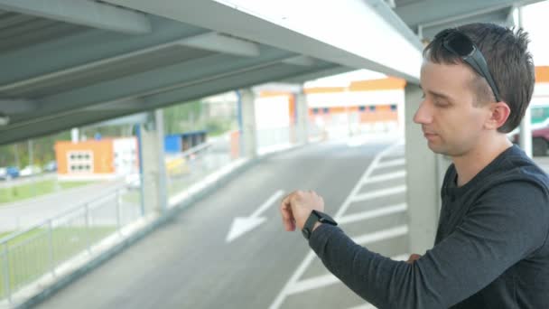 Retrato al aire libre del joven moderno con reloj inteligente en la calle. El hombre de gafas con mochila sentado en el puente. Coches de tracción inferior — Vídeos de Stock