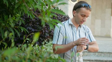 Modern genç adam akıllı saati olan açık portresi. güneş gözlüğü ve ceket. Çok yeşil bitkiler etrafında