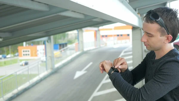 Outdoor portrait of modern young man with smart watch in the street. The  in glasses  backpack sitting on the bridge. Bottom drive cars