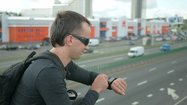 Outdoor portrait of modern young man with smart watch in the street. The  in glasses  backpack sitting on the bridge. Bottom drive cars