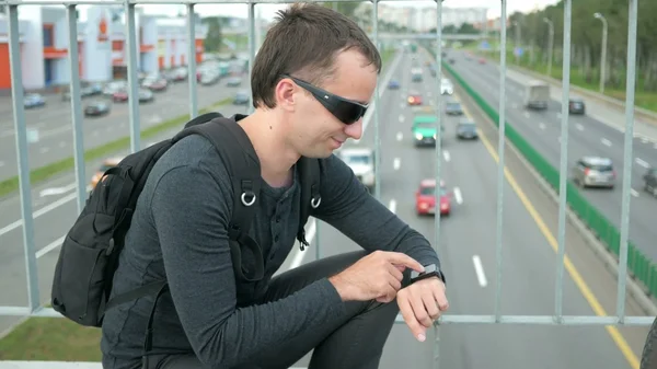 Outdoor portrait of modern young man with smart watch in the street. The in glasses backpack sitting on the bridge. Bottom drive cars