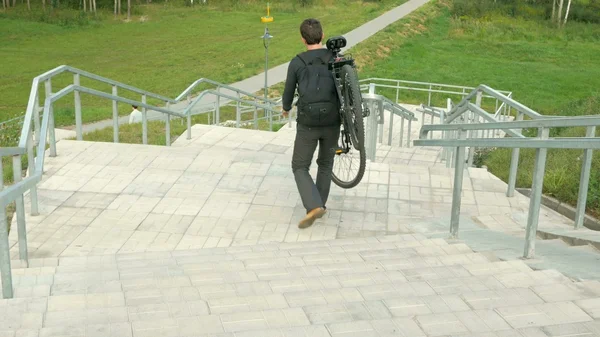 young man carries  bicycle through the long stairs. Against the background of green field. Clouds changing light