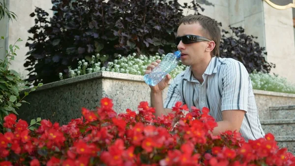 Thirsty man having a break drinking  bottle of water.  in sunglasses and jacket. Near  lot  red flowers