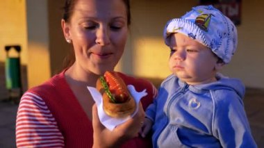 Gün batımında sosisli sandviç yeme güzel bebek ile anne. Beyaz kap ve mavi bisiklet, bir çocuk annesi ile bir sandviç yeme. Bir kafe ile bina arka planı.