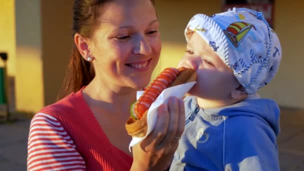 Matka z dzieckiem piękne jedzenie hot dogi o zachodzie słońca. Chłopiec w cap biały i niebieski rower, jeść kanapkę z matką. Na tle budynku z kawiarnią. — Wideo stockowe