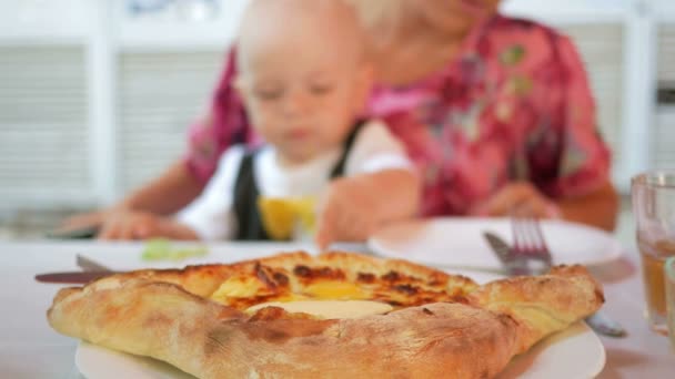 Mamá y el bebé en un hermoso vestido con mariposa comen en un café en la calle. Galleta de queso al horno con huevo y mantequilla en primer plano — Vídeos de Stock