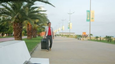 Genç bir turist büyük bir bavulla şehir parkının etrafında dolanıyor. Durup etrafa bakıyor. Beyaz tişörtlü bir adam..