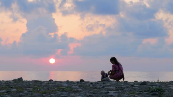 Família Schislivaya: mãe, filhinho na praia rochosa do mar durante o pôr do sol. Pai brincar e beijar bebê nos raios do sol vermelho e céu azul — Vídeo de Stock