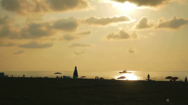 Tramonto sulla spiaggia del mare. Silhouette di persone sulla spiaggia. La barca sta navigando al sole — Video Stock