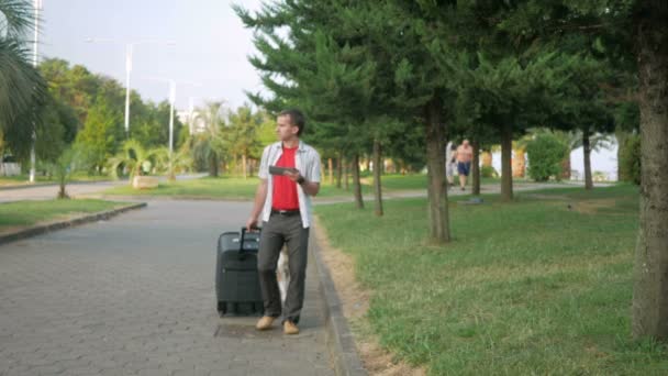 Jovem turista é com uma grande mala sobre rodas em torno do parque da cidade. Nas mãos de um tablet com um tablet e está procurando o caminho certo. Perto anda cão vadio . — Vídeo de Stock