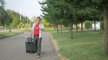 Genç bir turist büyük bir bavulla şehir parkının etrafında dolanıyor. Akıllı saatlerle mesaj geldi, adam durdu ve cevabı dikte etti. Yakınlarda başıboş bir köpek dolaşıyor.