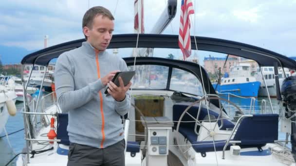 ヨットの近くに立っているハンサムな若者。彼はタブレット上のメッセージをチェックし、ソーシャルネットワークで友人に会います. — ストック動画