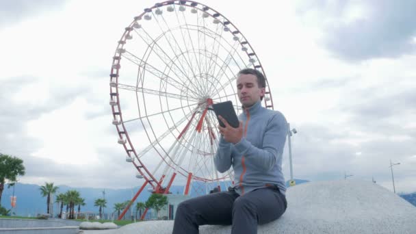 A young man checks the messages on the Tablet PC around the Ferris wheel. Early morning. The wheel does not spin. — Stock Video