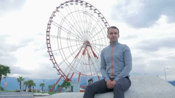 Young handsome man looking into the camera and smiles around the Ferris wheel. Early morning. The wheel does not spin. — Stock Video