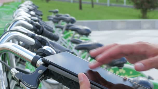 Un hombre comprueba los mensajes en el Tablet PC en un estacionamiento para bicicletas. Va a alquilar una bicicleta. — Vídeo de stock