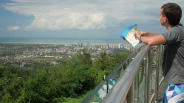 Yüksek katlı binalar ve sıradan evler ile modern bir şehir. Bir adam bir kağıt harita rotasında denetler. Orman ve deniz arkasında