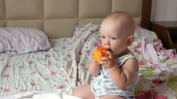 Un bebé guapo comiendo un melocotón en un desayuno en la cama. Le arranca un pedazo y se lo mete en la boca. — Vídeo de stock