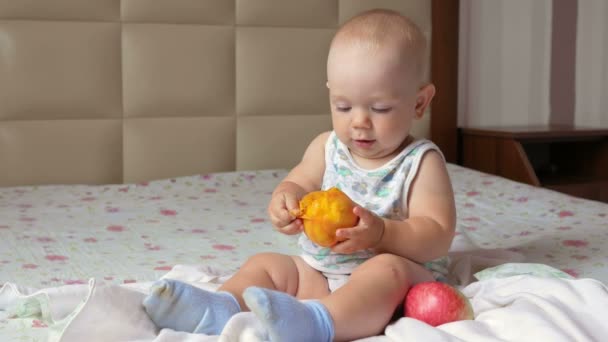 Ένα όμορφο μωρό τρώει ένα ροδάκινο σε ένα πρωινό στο κρεβάτι. Αυτός δάκρυα από ένα κομμάτι και βάζει στο στόμα του — Αρχείο Βίντεο